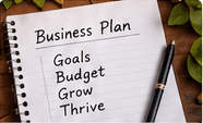A spiral notebook headed "Business Plan", listing goals, budget, grow and thrive, with a pen laid across it.